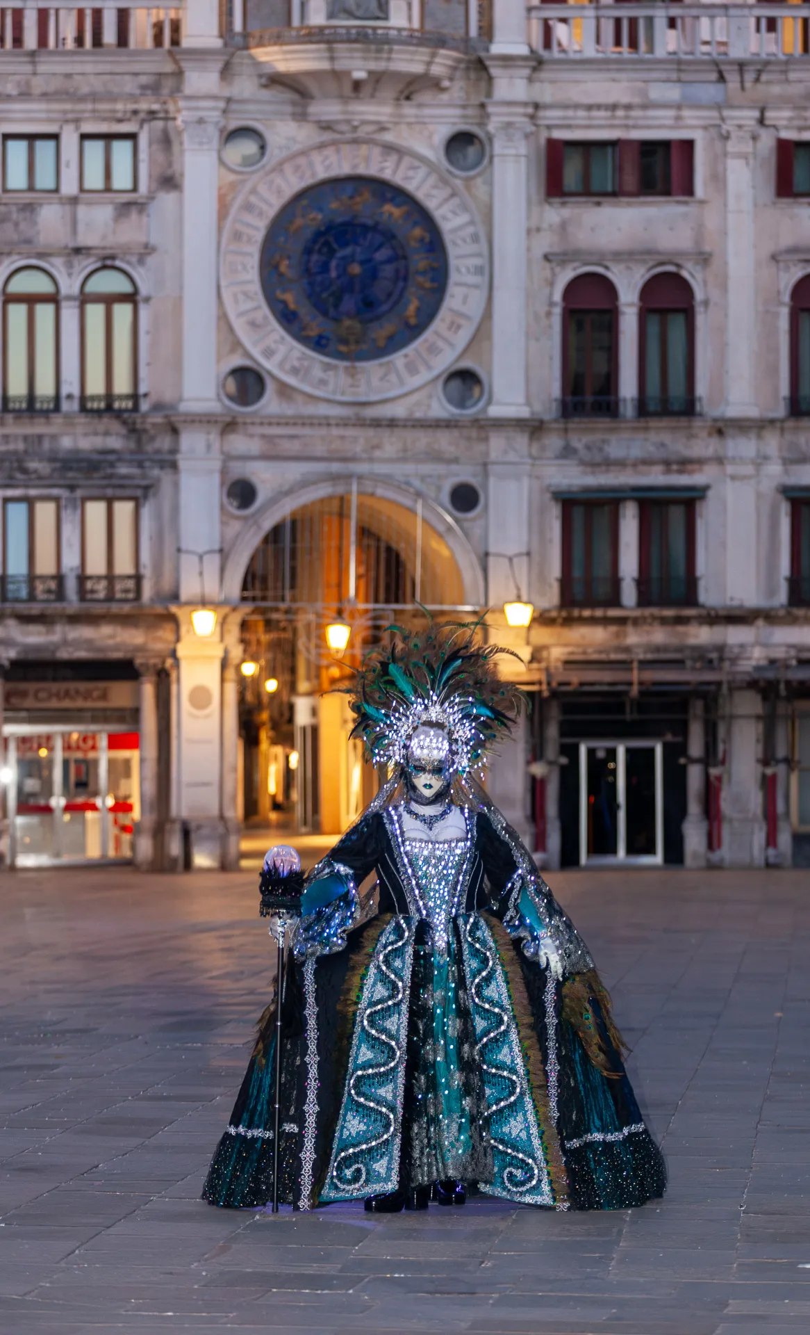 Carnevale di Venezia 2025 all'alba: una figura mascherata illuminata si staglia davanti alla Torre dell'Orologio, catturando la magia del momento.
