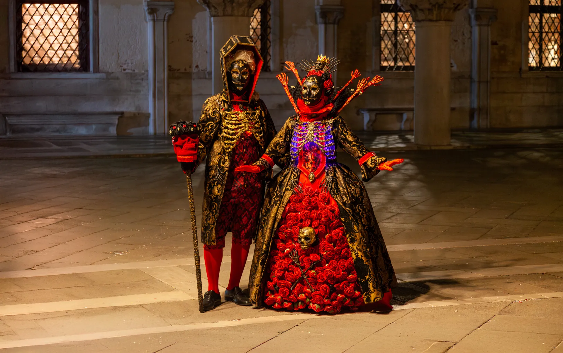 Coppia in maschera barocca con dettagli dorati e rossi sotto i portici di Palazzo Ducale.