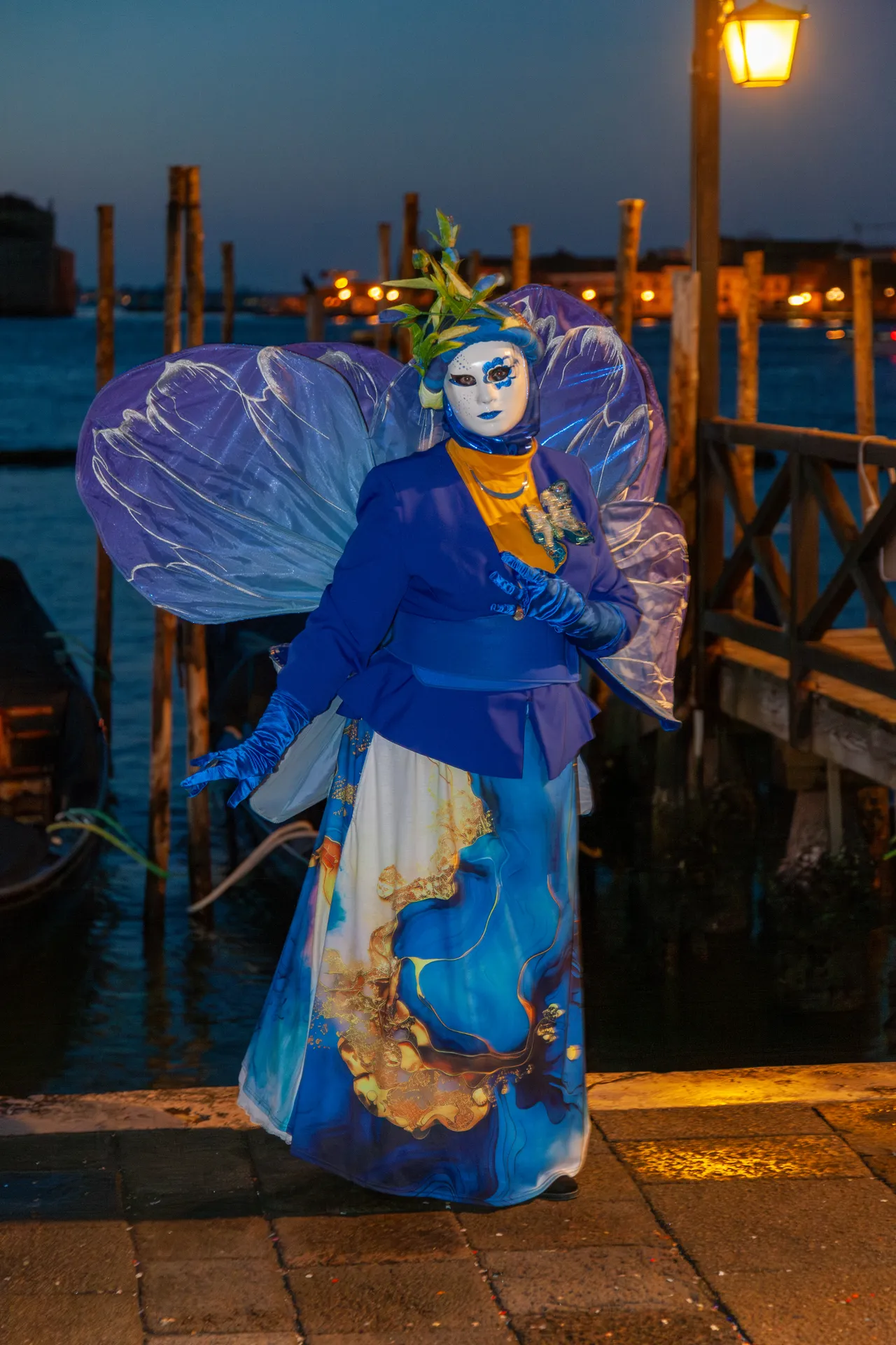 Dama con ali azzurre e maschera bianca posa sulla riva veneziana al mattino.