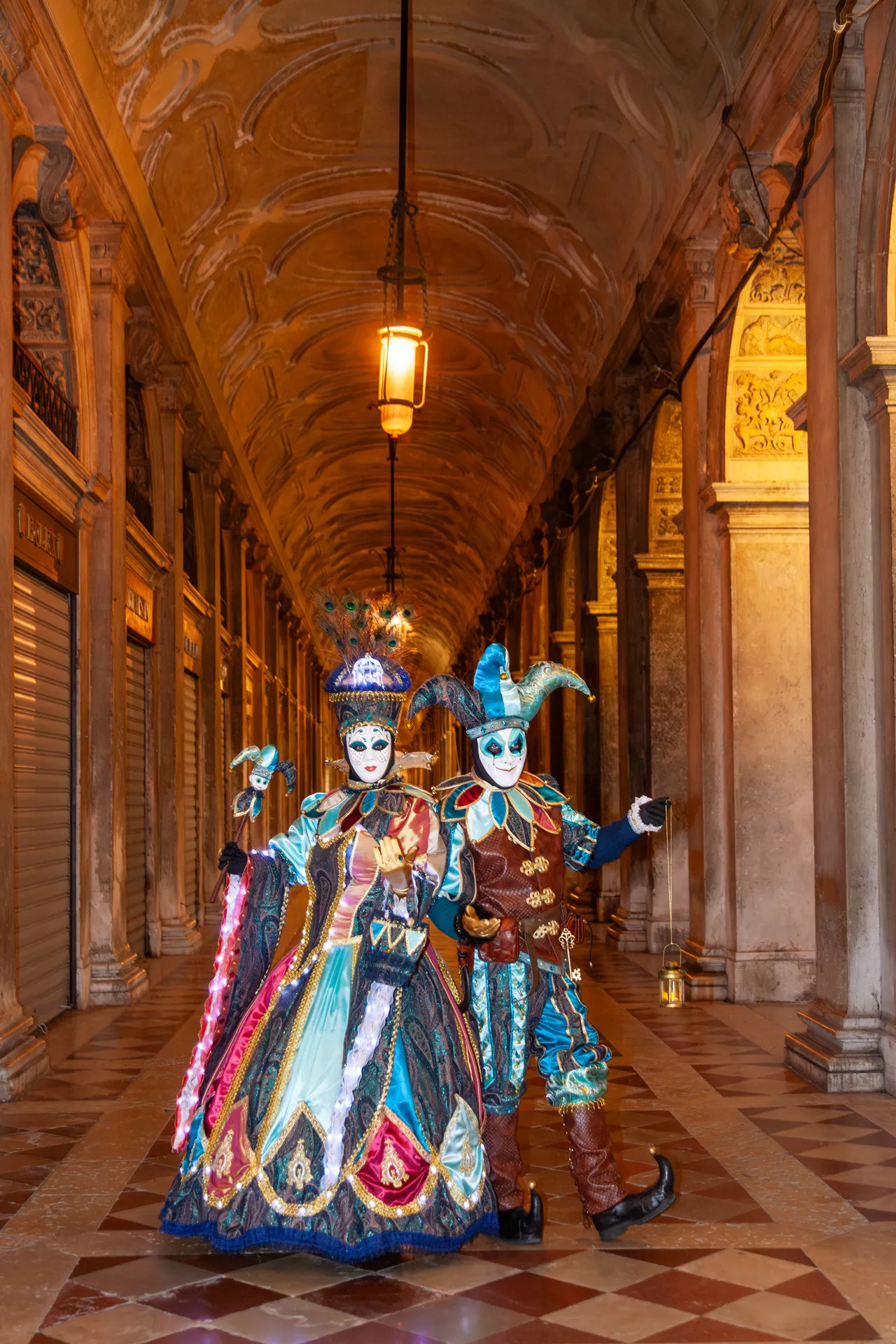 Foto scattata durante il Carnevale di Venezia 2025, con due maschere in abiti sfarzosi sotto i portici  delle Procuratie Nuove, illuminati all'alba.