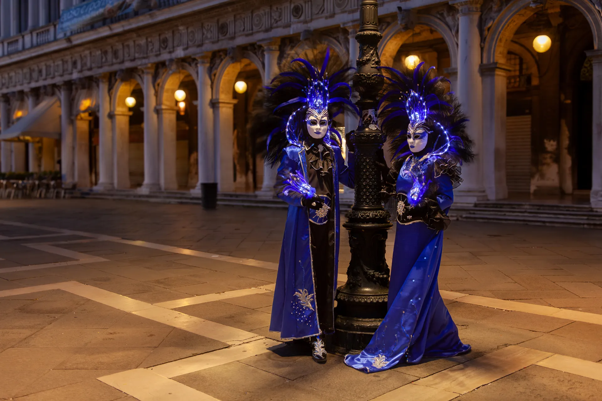 Foto realizzata all'alba al Carnevale di Venezia 2025, immortalando due maschere in abiti blu decorati con piume illuminate.