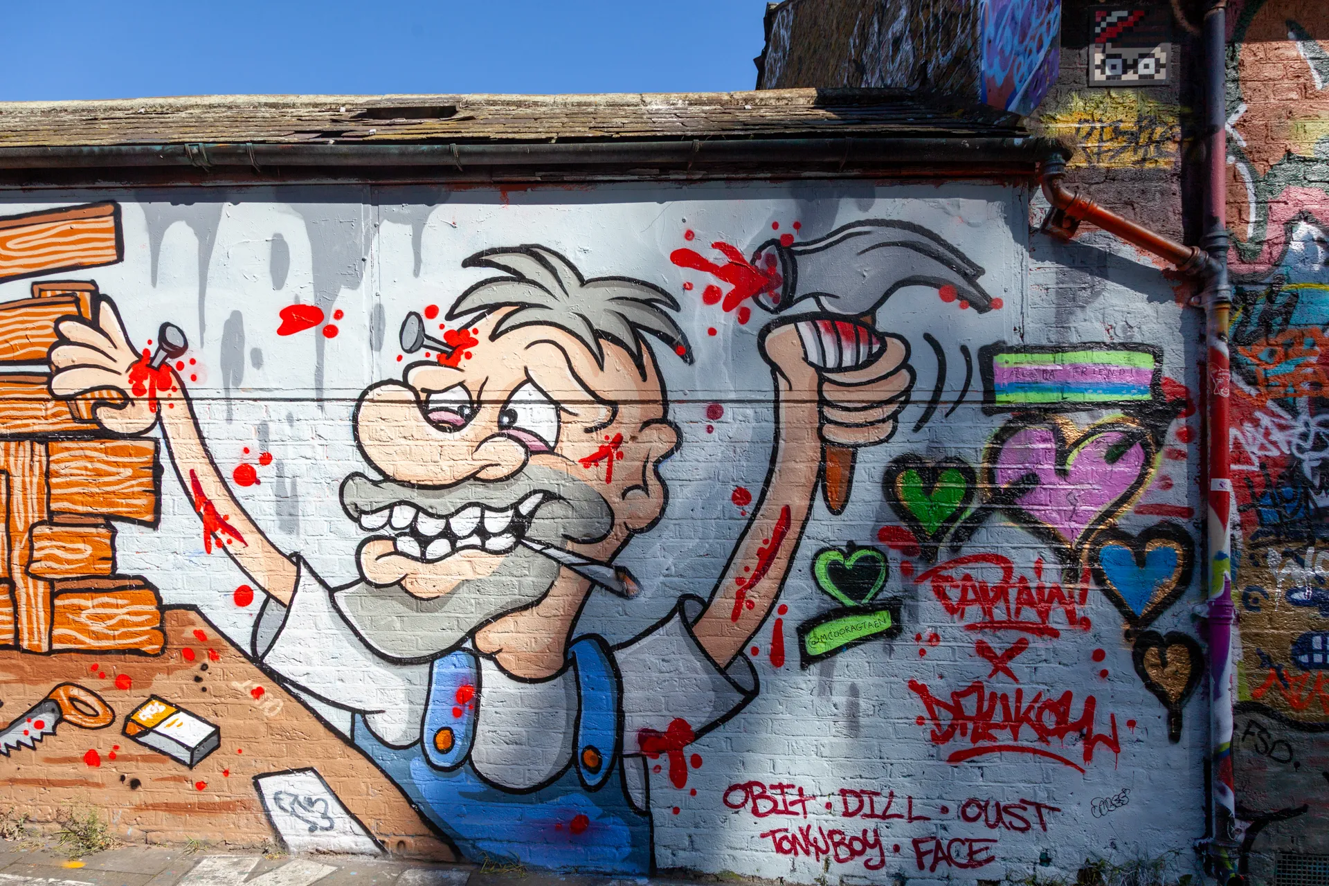 Murale grottesco di un carpentiere anziano a Brick Lane, firmato da un collettivo di artisti (Obit, Dill, Oust, Tonyboy, Face). Ironia, colori forti e atmosfera ribelle nel cuore di Londra. Muro laterale vicino a Bacon Street, Brick Lane