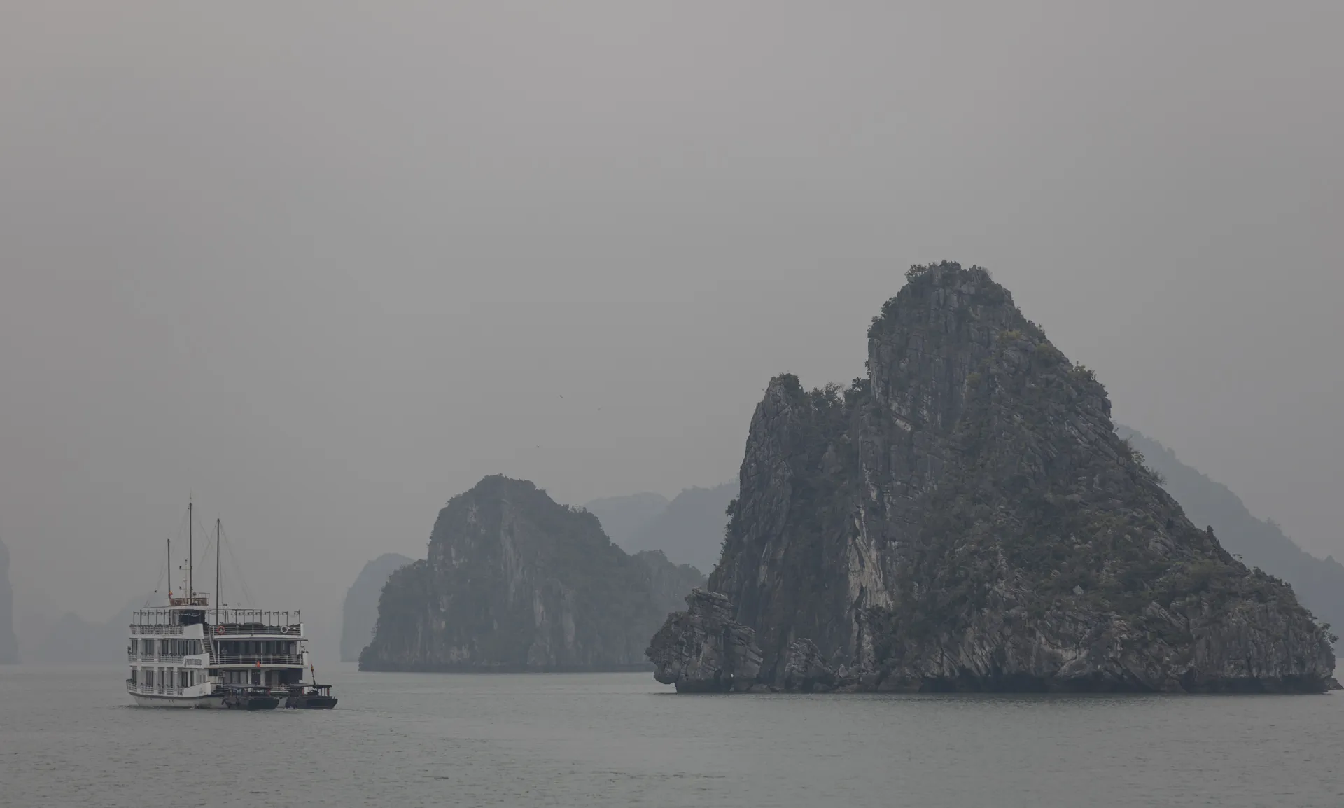 La baia di Ha Long avvolta dalla foschia, con profili carsici e una barca da crociera sullo sfondo.